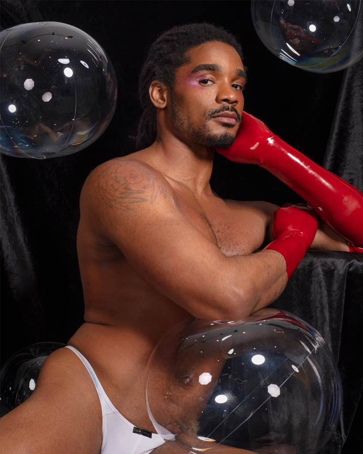Khalyle surrounded by transparent bubbles, posed leaning with his body outstretched in only a white thong and red latex gloves looking at the camera.